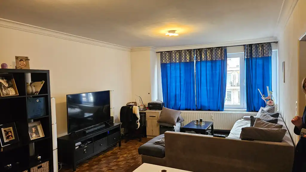Salon avant rénovation avec canapé gris, grande fenêtre voilée de rideaux bleus et meuble TV noir