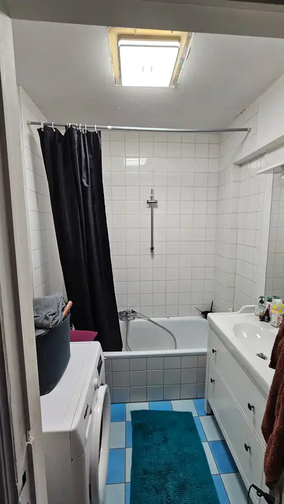 Salle de bain ancienne avec baignoire, carrelage blanc, machine à laver et sol en carrelage bleu et blanc.