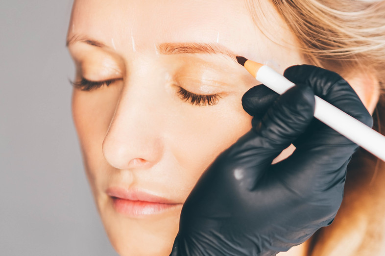 woman-makeup-master-applies-color-on-eyebrow-of-attractive-client-with-pencil-near-white-w