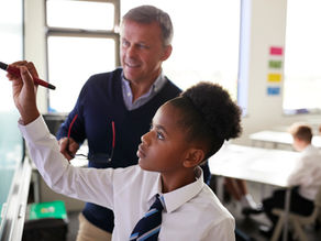Student in uniform writes on a whiteboard, guided by a teacher in a classroom. Bright light from windows, engaged learning atmosphere.
