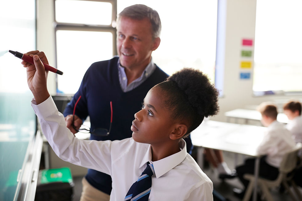 Student in uniform writes on a whiteboard, guided by a teacher in a classroom. Bright light from windows, engaged learning atmosphere.