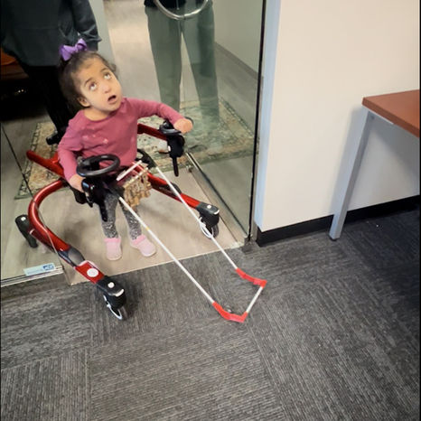 A 4-year-old girl using a gait trainer and wearing a belt cane walks through a doorway