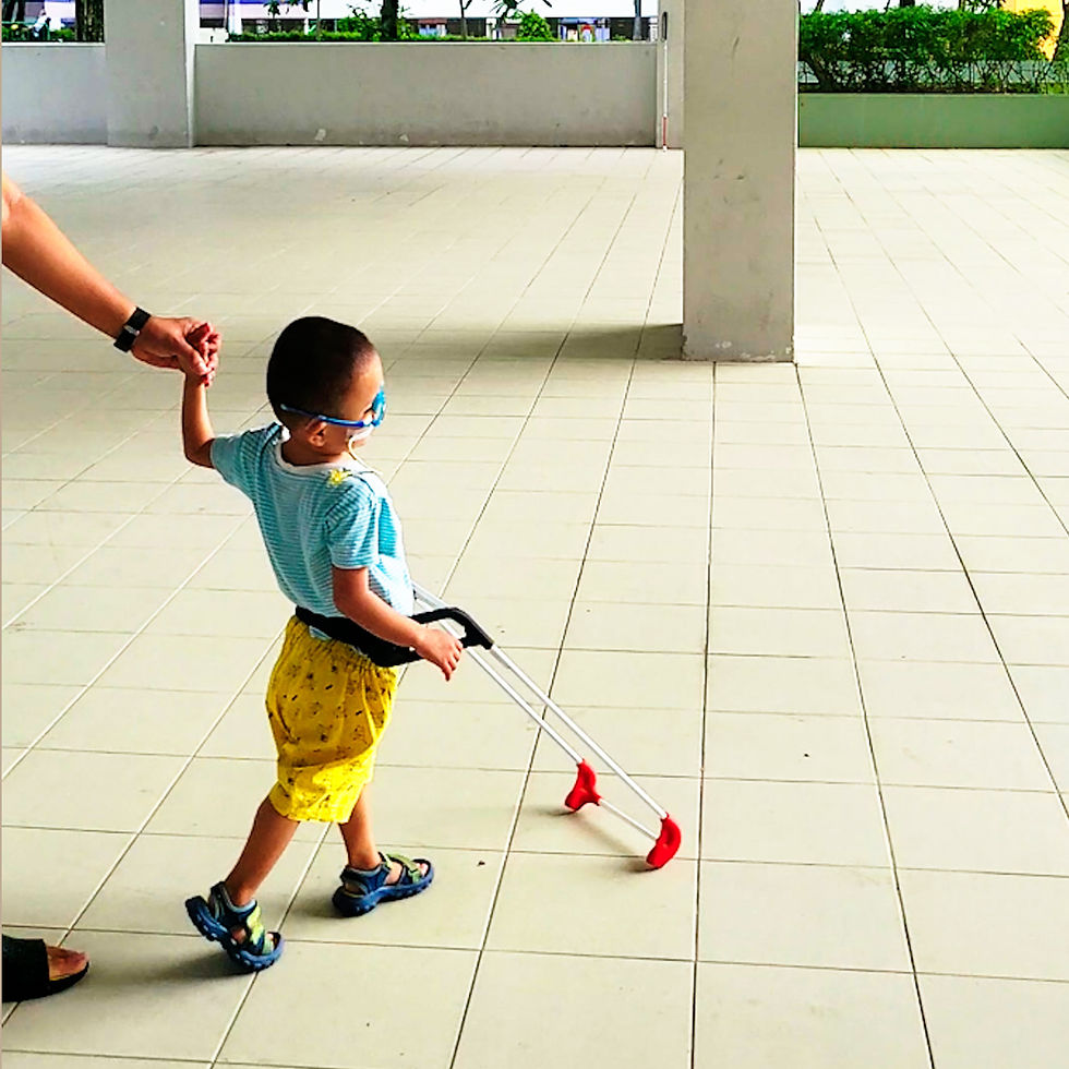 Child in blue shirt and yellow shorts walks wearing a belt cane while holding an adult’s hand and leading the way in a bright tiled corridor.