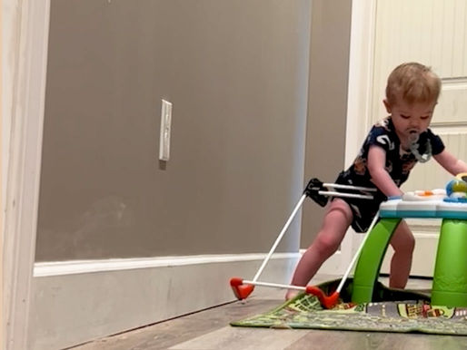 Toddler wearing a Belt Cane on a playmat pushing a standing toy, pacifier in mouth, in a hallway with beige walls and wood flooring, appearing focused.