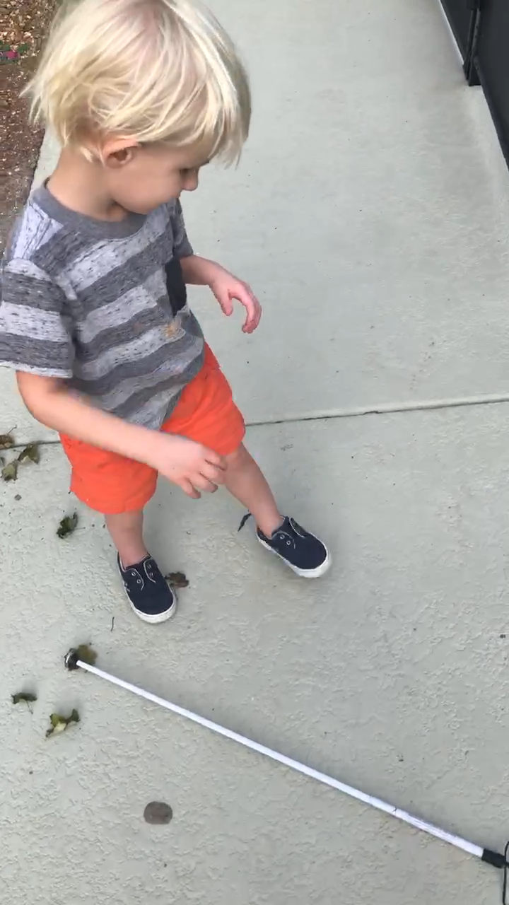 a 3-year-old boy stands on the sidewalk his long cane lies on the ground.
