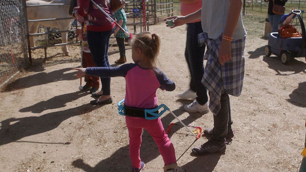 Young girl wearing a Belt Cane at a farm pen while adults and children stand nearby at a sunny petting zoo.