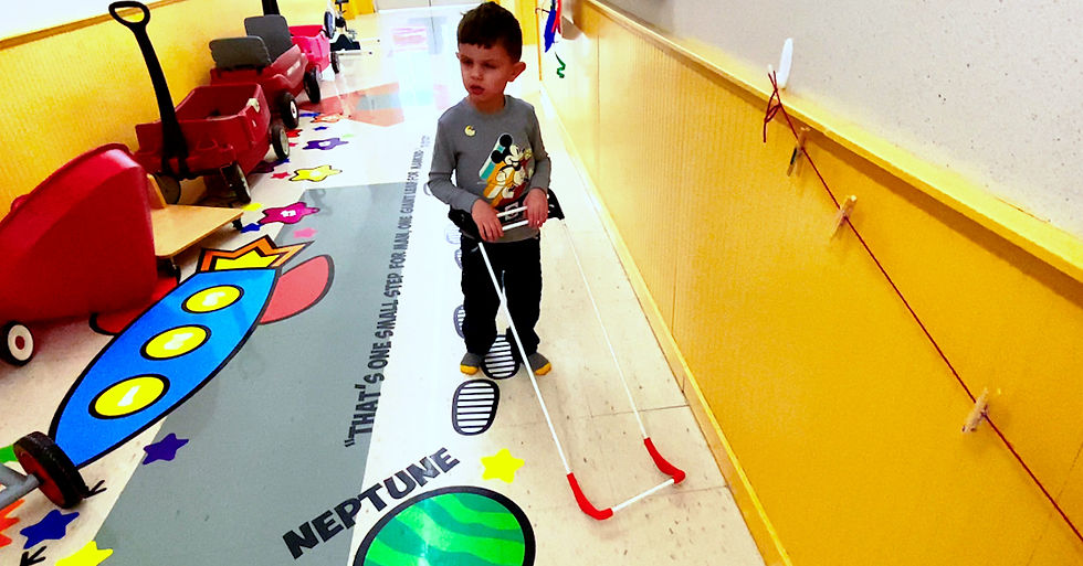 Young boy in a colorful hallway wearing a red and white belt cane. Floor has planets and footprints with text: "That's one small step..."