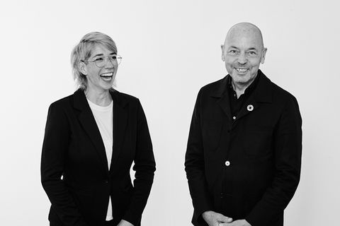 Team Porträt in Schwarz weiß. Frau mit kurzem Haar, Brille und dunklem Blazer steht leicht schräg zur Kamera und schaut lachend zur Seite. Man mit Glatze und schwarzer Jacke steht neben ihr frontal zur Kamera und lacht zur Kamera.