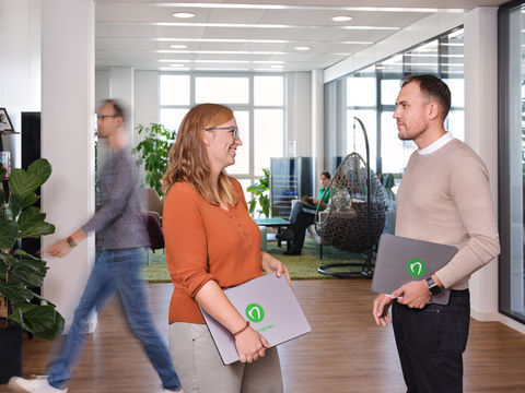 Zwei Mitarbeiter stehen in einem offenen Büro und unterhalten sich. Sie haben Laptops mit dem firmen Logo in der Hand.
