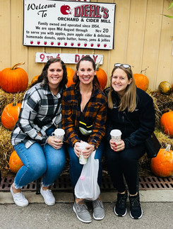 Three women sit on haybales surrounded by pumpkins underneath signage for Diehl's Orchard and Cider Mill