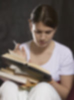 Brown-haired Girl wearing white sitting with a book and pencil appearing to take notes