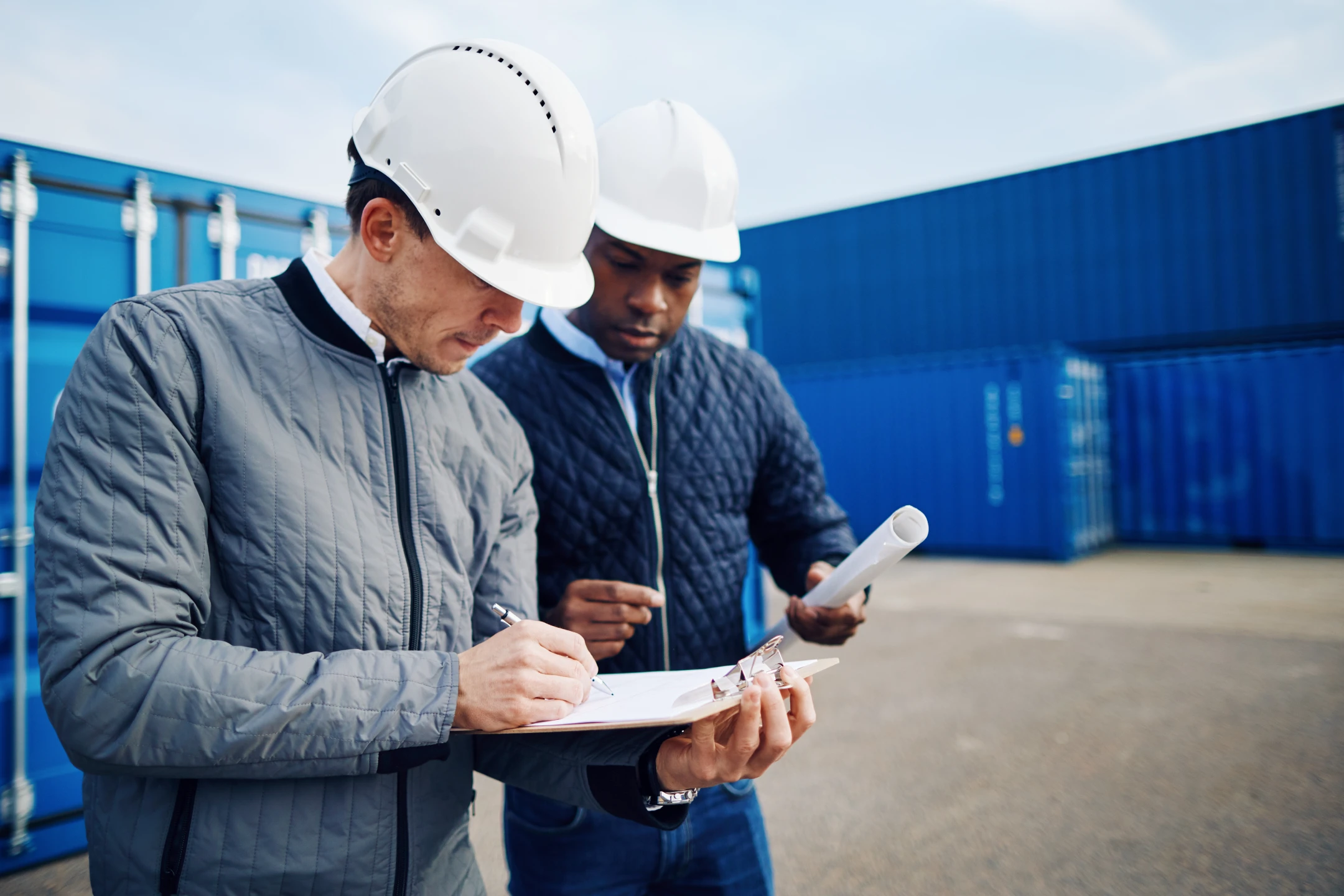 Logistics personnel wearing hard hats looking at clipboard with blue shipping contatiners in the background