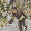 A person arranging a long outdoor table decorated with white flowers and greenery, holding a yellow notepad while adjusting t