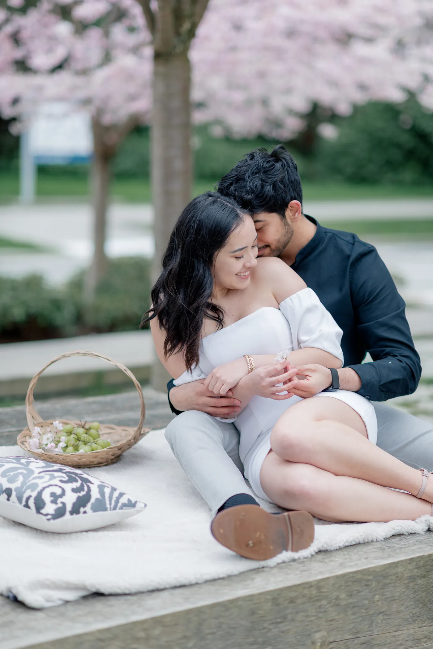 A soft and intimate picnic scene under the cherry blossoms. The couple enjoys each other's presence while soaking in the beauty of the blooming trees.