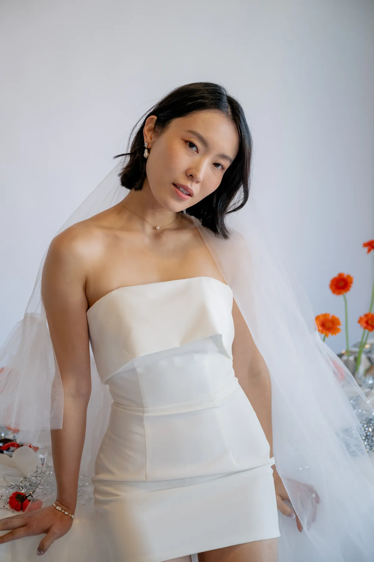 A bride wearing a sleek white mini dress and veil, holding a bold bouquet of orange gerbera daisies and purple hydrangeas, in an industrial wedding setting.