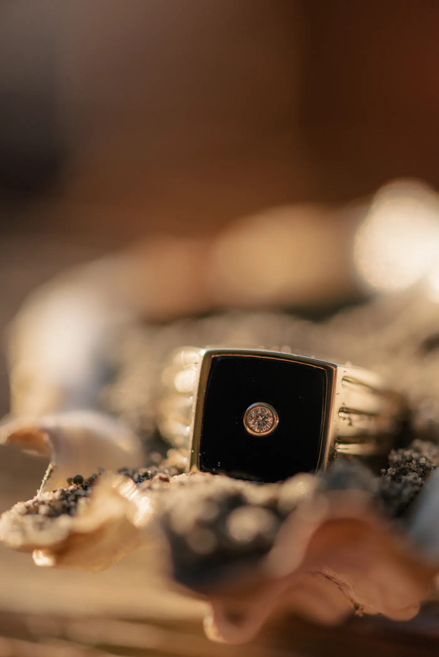 A unique gold and black engagement ring with a diamond centerpiece, placed on a seashell with sand. A modern and stylish ring choice for a queer proposal in Canada, captured in warm golden light.