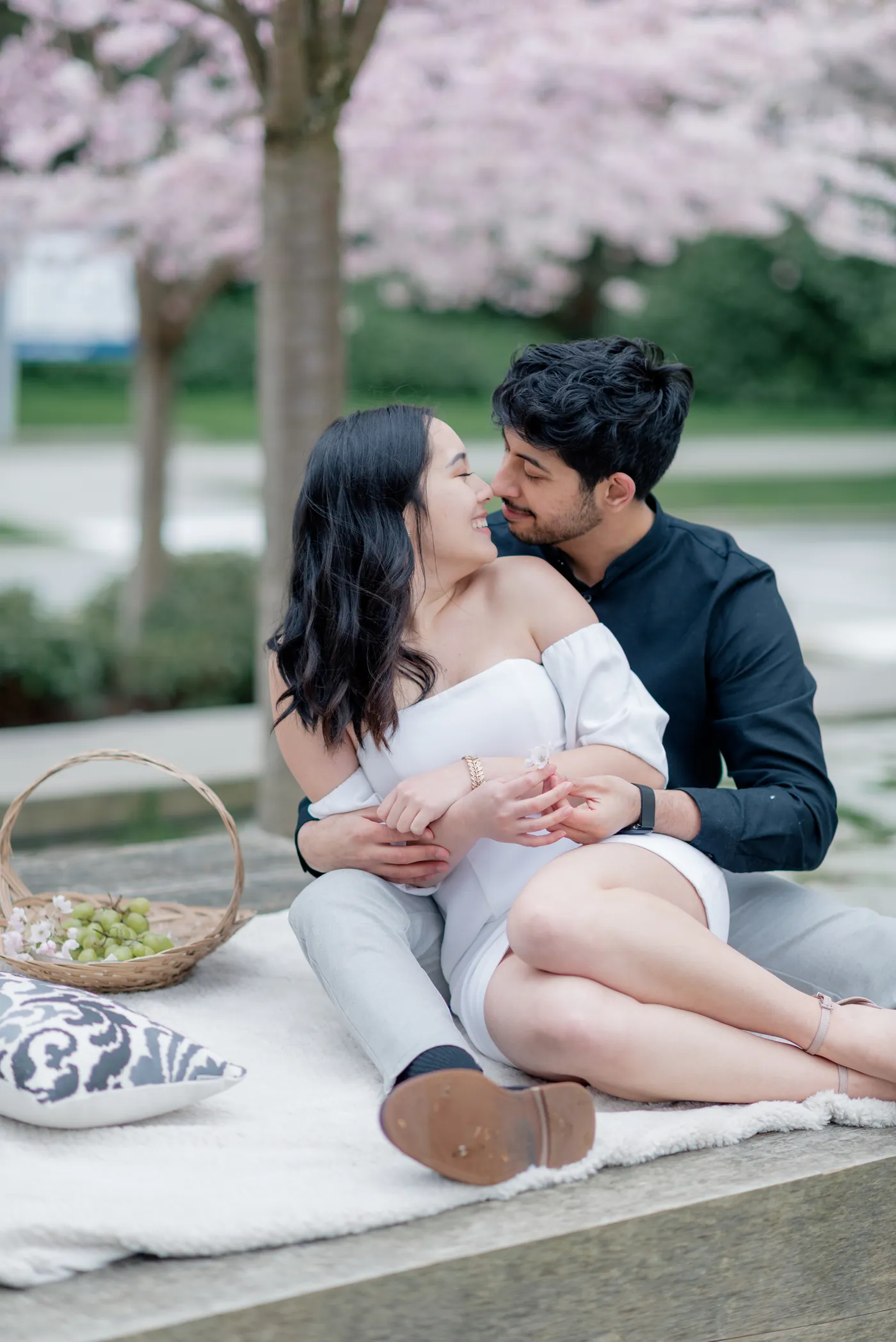 A soft and intimate picnic scene under the cherry blossoms. The couple enjoys each other's presence while soaking in the beauty of the blooming trees.