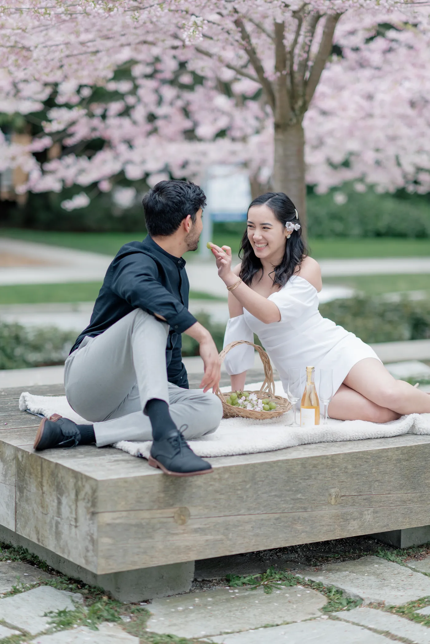 A couple enjoys a spring picnic, reaching into a basket filled with grapes and blossoms while sharing a peaceful moment together.