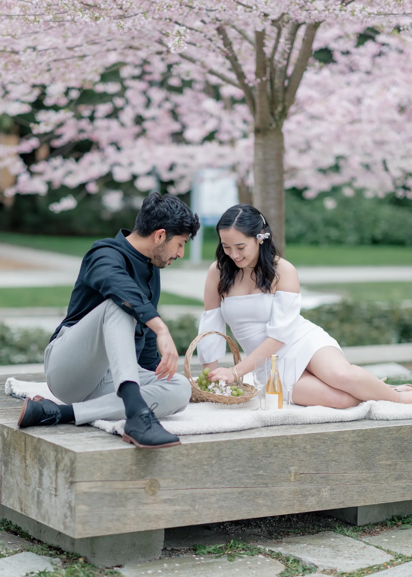A couple enjoys a spring picnic, reaching into a basket filled with grapes and blossoms while sharing a peaceful moment together.