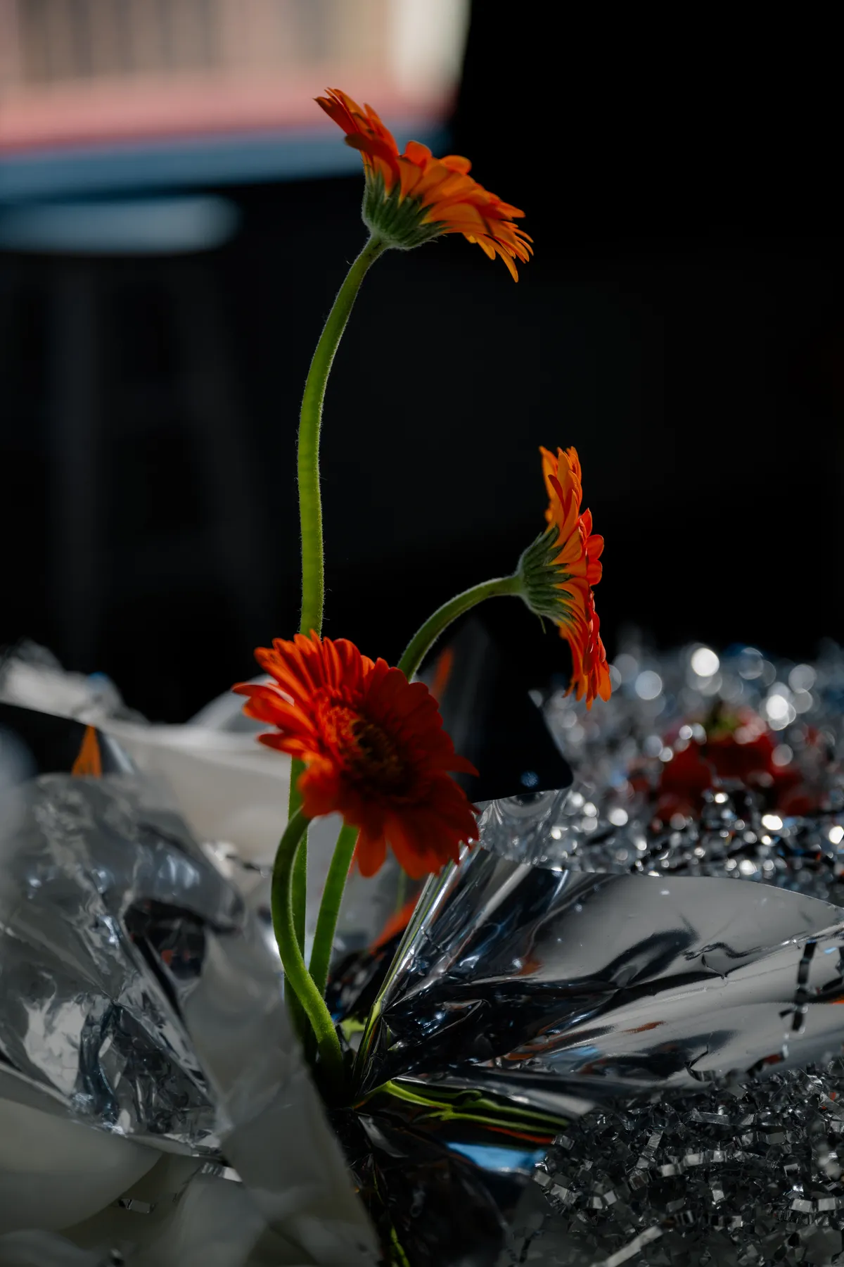 A close-up of a moody reception table with crystal glassware, bold orange gerbera daisies, shimmering silver details, and a white tablecloth.
