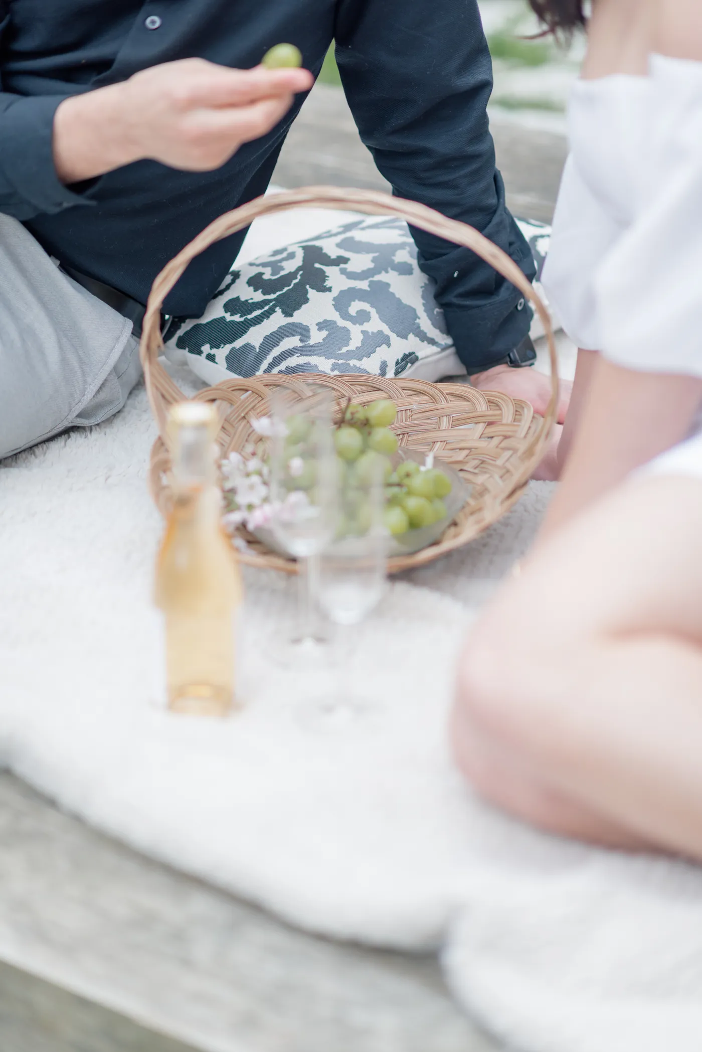 A couple enjoys a spring picnic, reaching into a basket filled with grapes and blossoms while sharing a peaceful moment together.