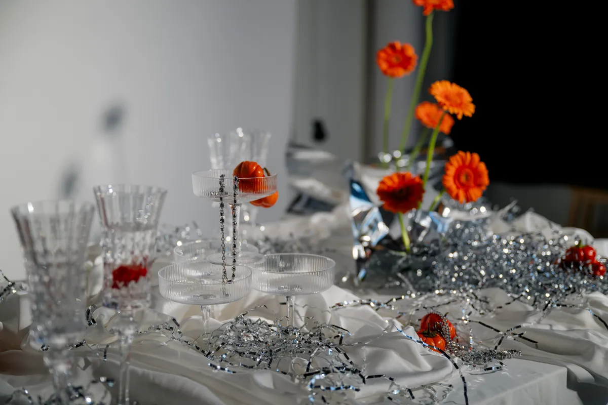 A close-up of a moody reception table with crystal glassware, bold orange gerbera daisies, shimmering silver details, and a white tablecloth.