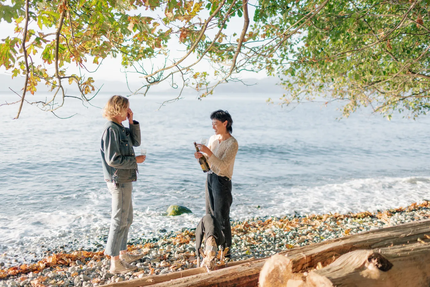 Newly engaged same-sex couple celebrating by the ocean with champagne. A candid and joyful moment during a post-proposal photography session on the Canadian coast.