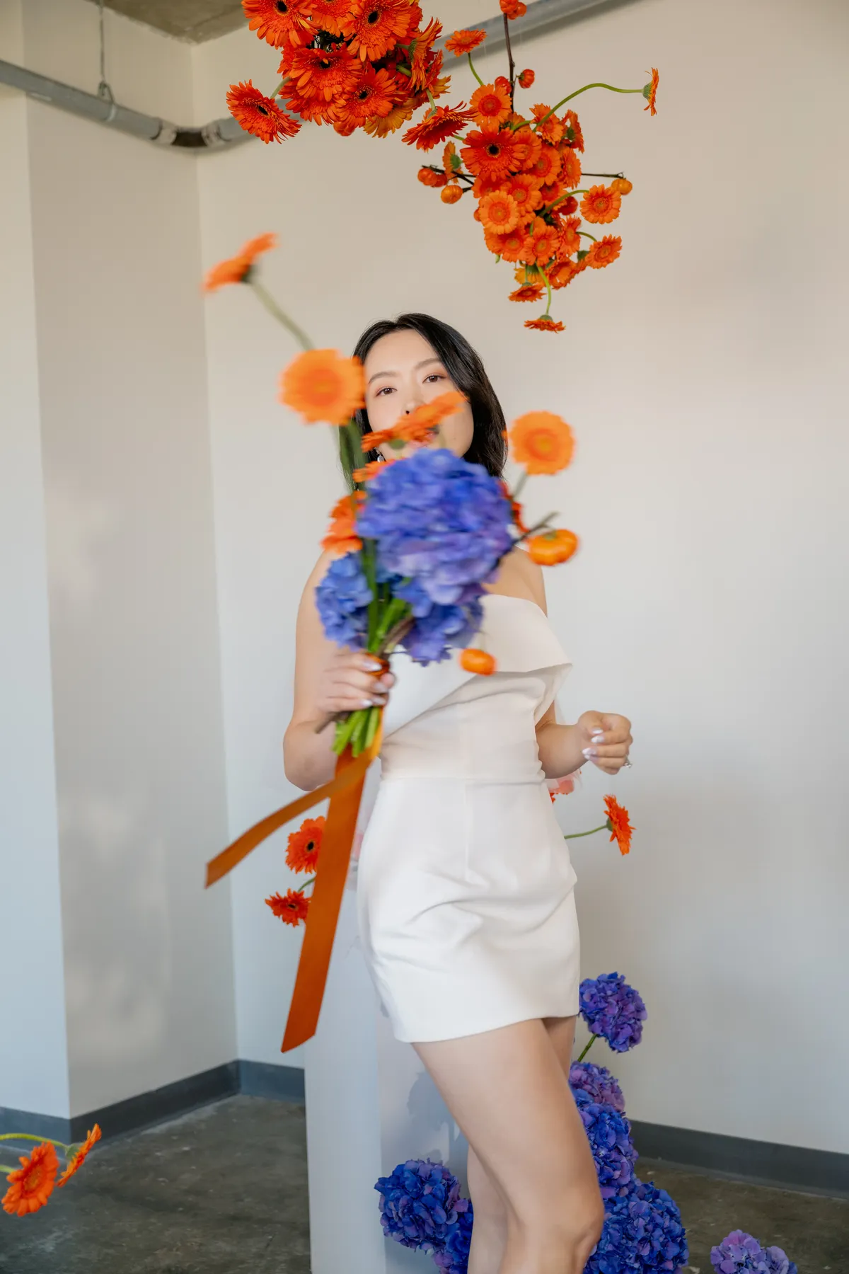 A bride wearing a sleek white mini dress and veil, holding a bold bouquet of orange gerbera daisies and purple hydrangeas, in an industrial wedding setting.