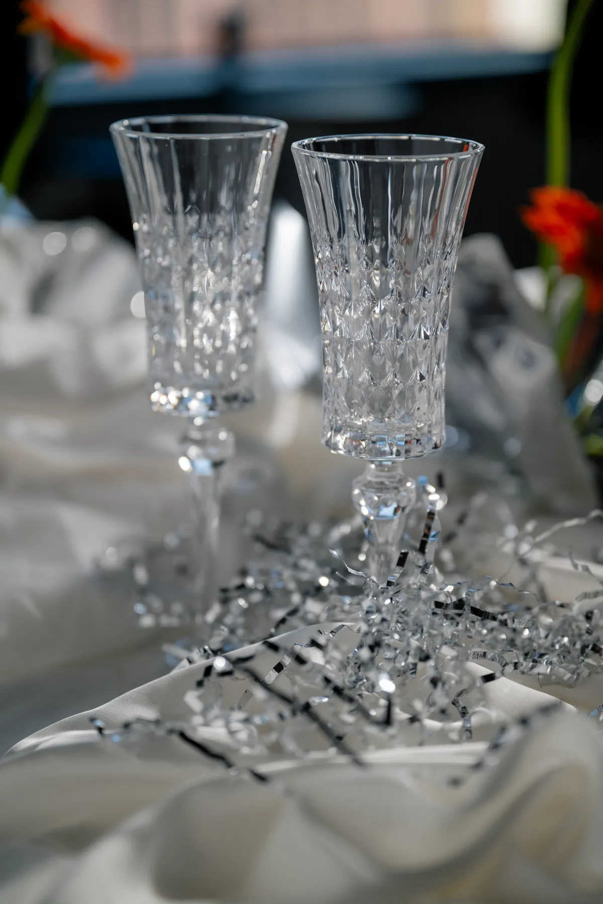 A close-up of a moody reception table with crystal glassware, bold orange gerbera daisies, shimmering silver details, and a white tablecloth.