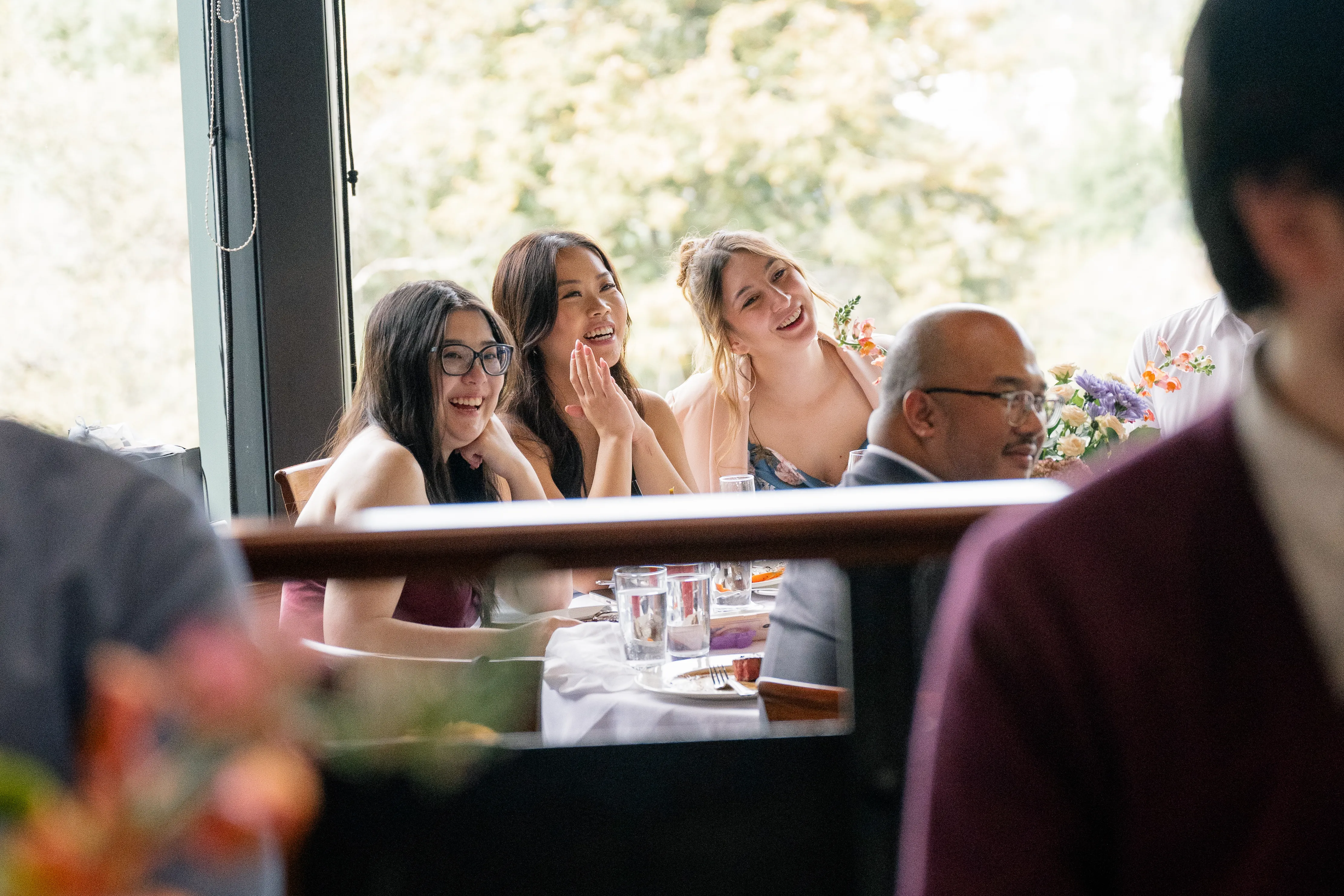 Three guests laughing and smiling as they listen to a speech during the wedding reception.