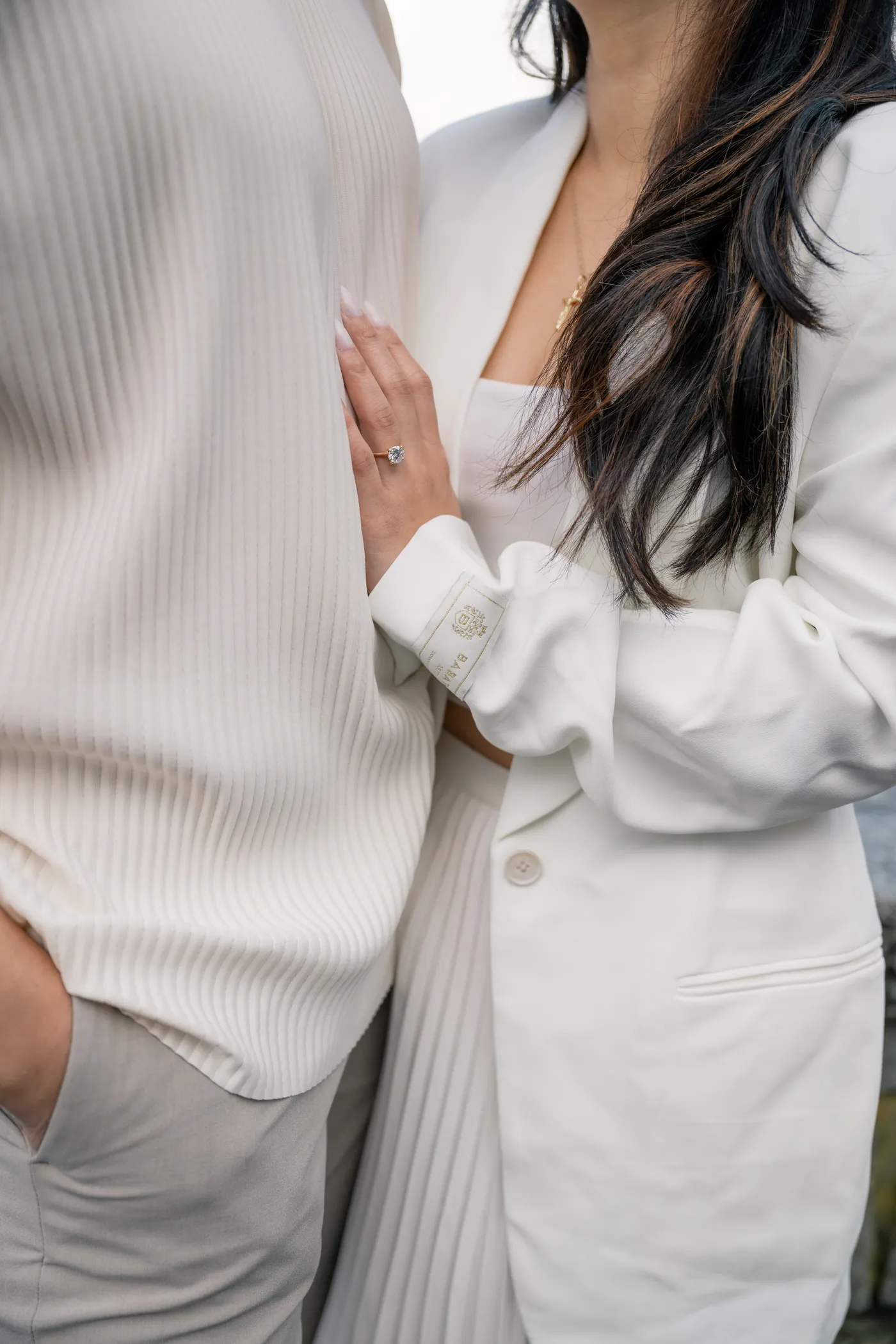 A close-up of a woman's hand resting on her partner's chest, showcasing an engagement ring. Both are dressed in elegant neutral tones.