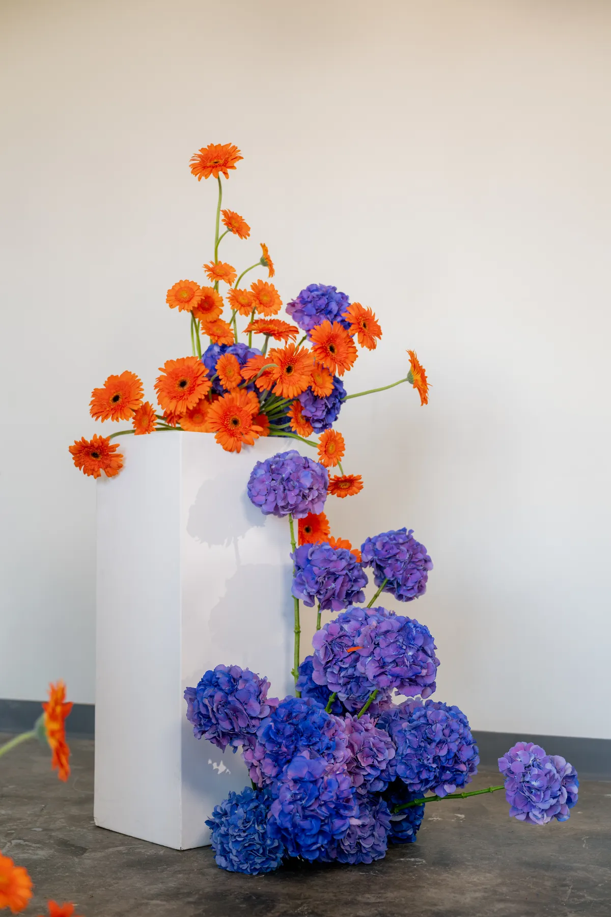 Suspended orange flowers and purple hydrangeas on modern white pedestals in a minimalist industrial venue, showcasing bold wedding decor.