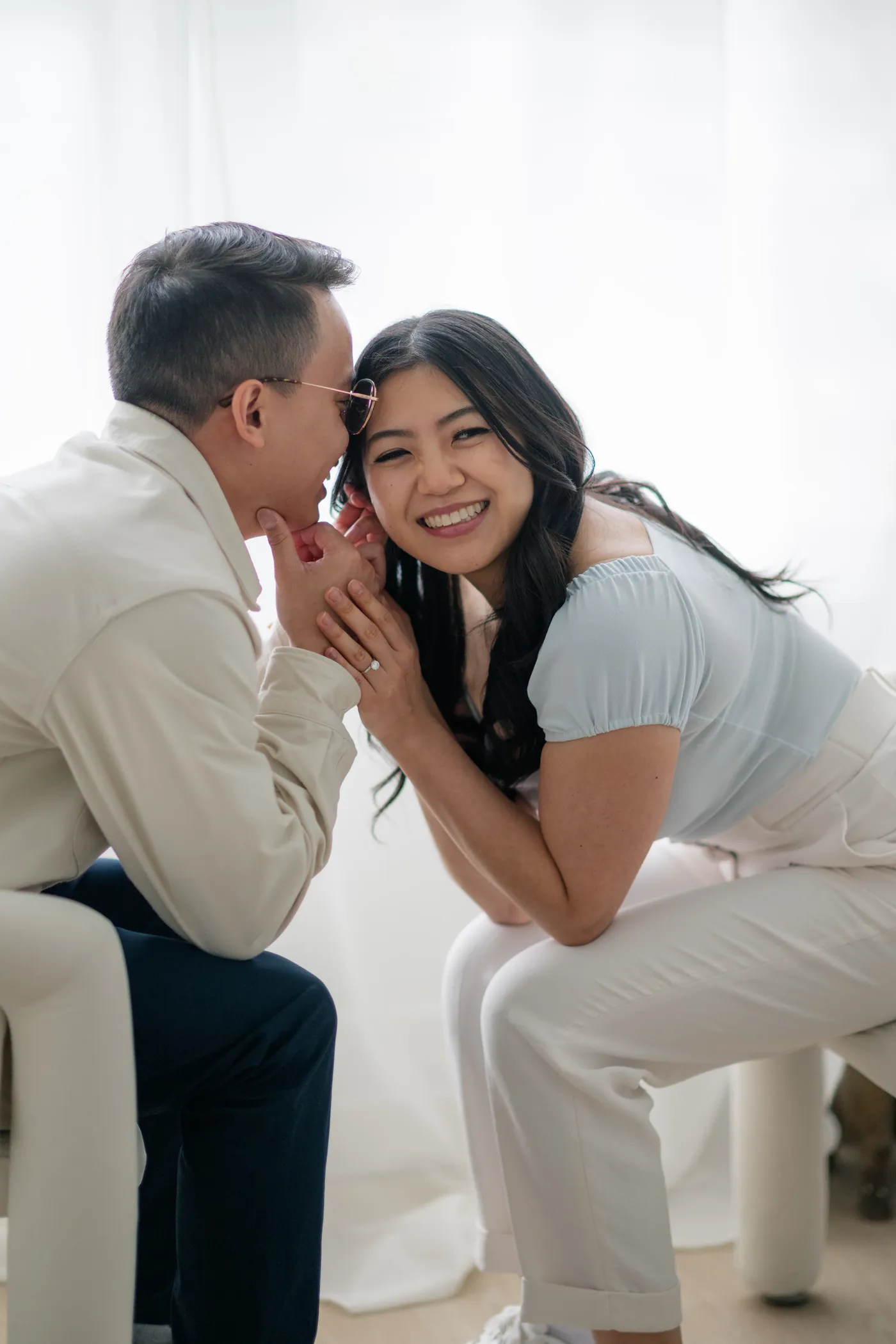 Close-up of engaged couple holding hands with soft natural light streaming in through large studio windows.