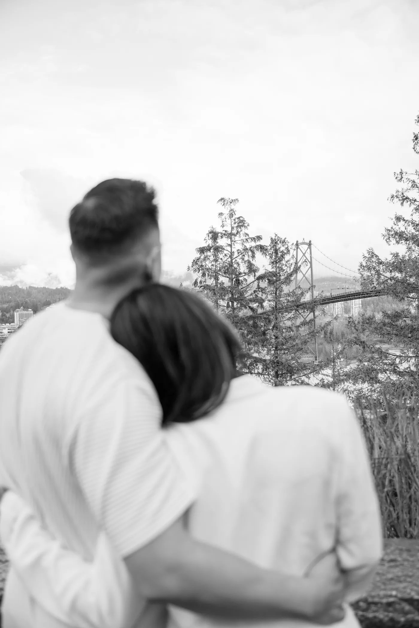 A couple embraces in front of a scenic overlook, smiling at each other as the city skyline and mountains stretch behind them.