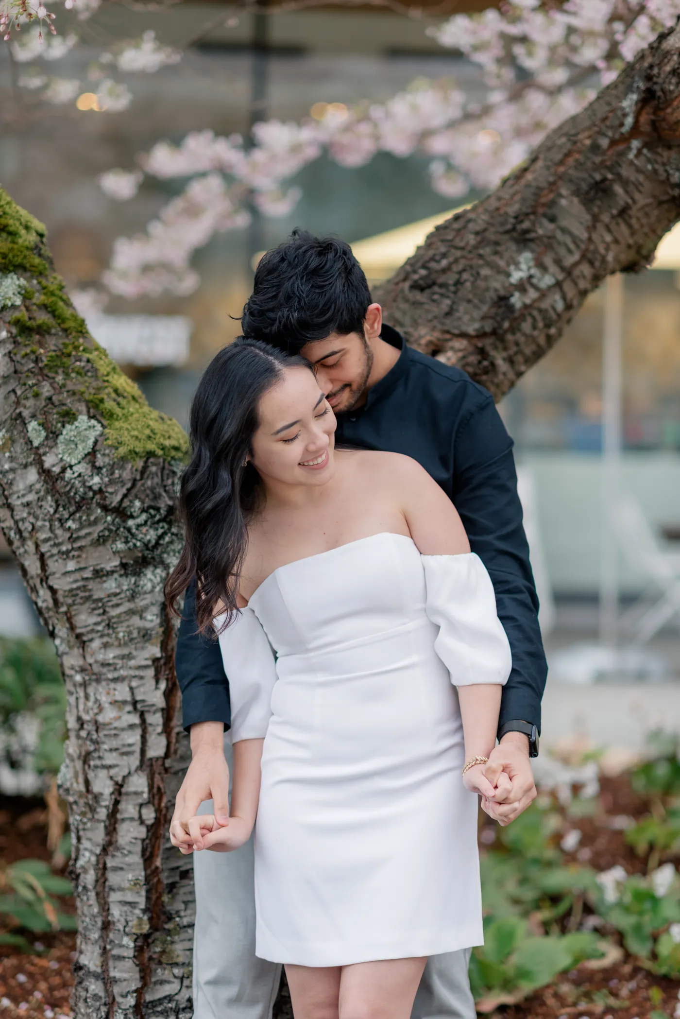 A dreamy moment of love as a couple stands under a cherry blossom tree, holding hands and gazing into each other’s eyes. The soft pink petals frame their embrace.