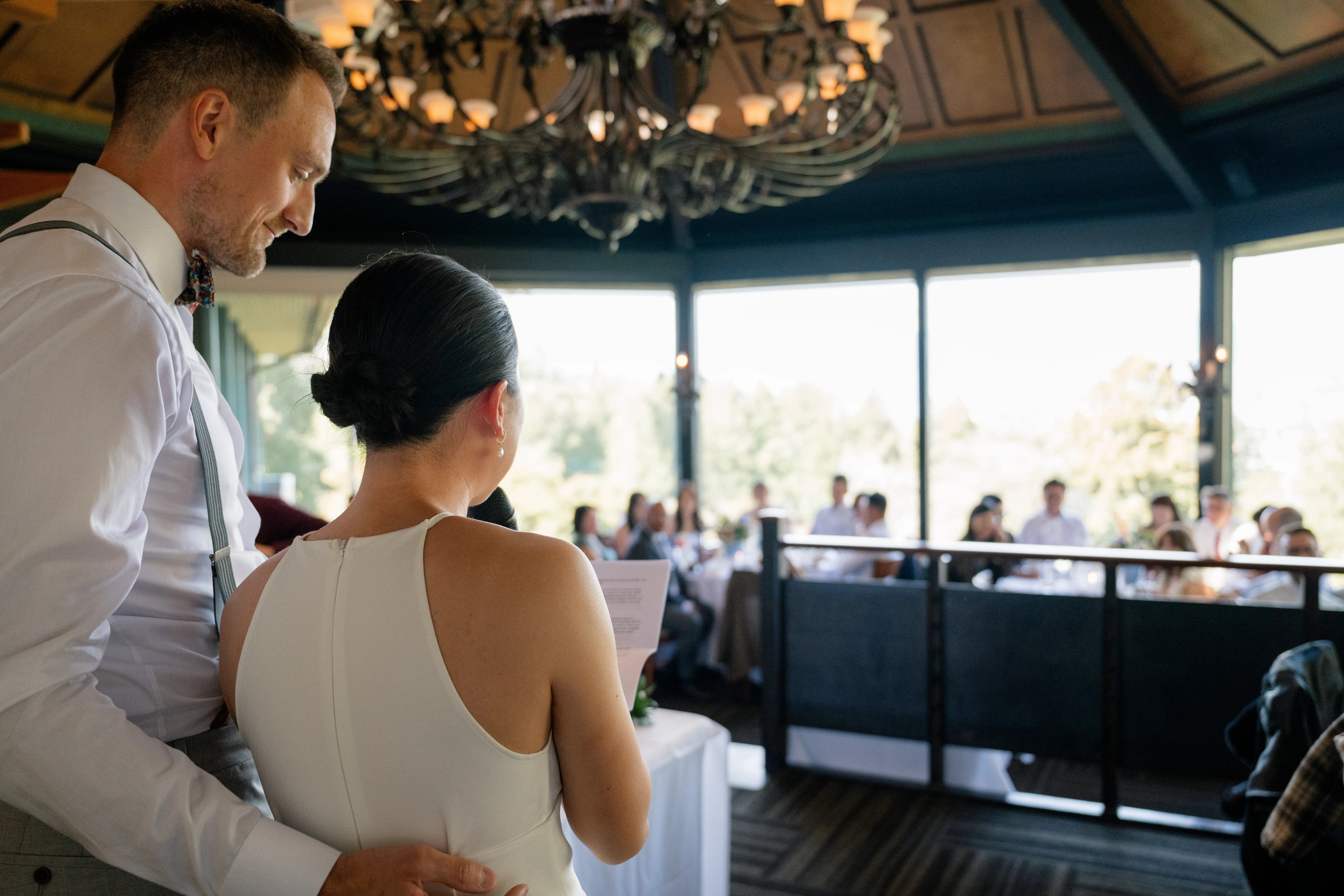 Bride speaking at the wedding reception, facing her guests, with chandeliers and large windows framing the elegant venue.