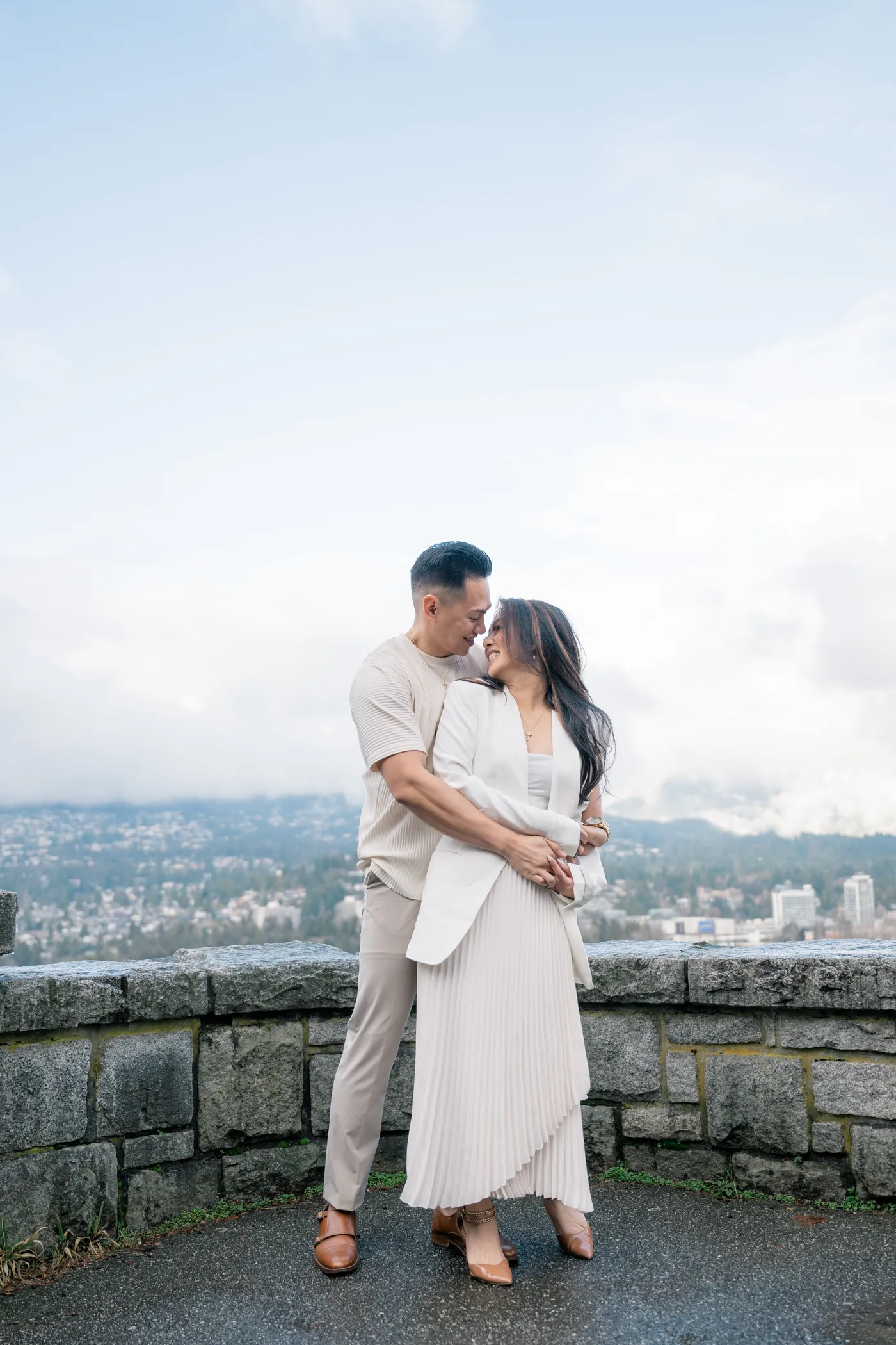 A couple joyfully dances on a stone platform at Prospect Point, with the Vancouver skyline and ocean in the background, creating a playful engagement scene.