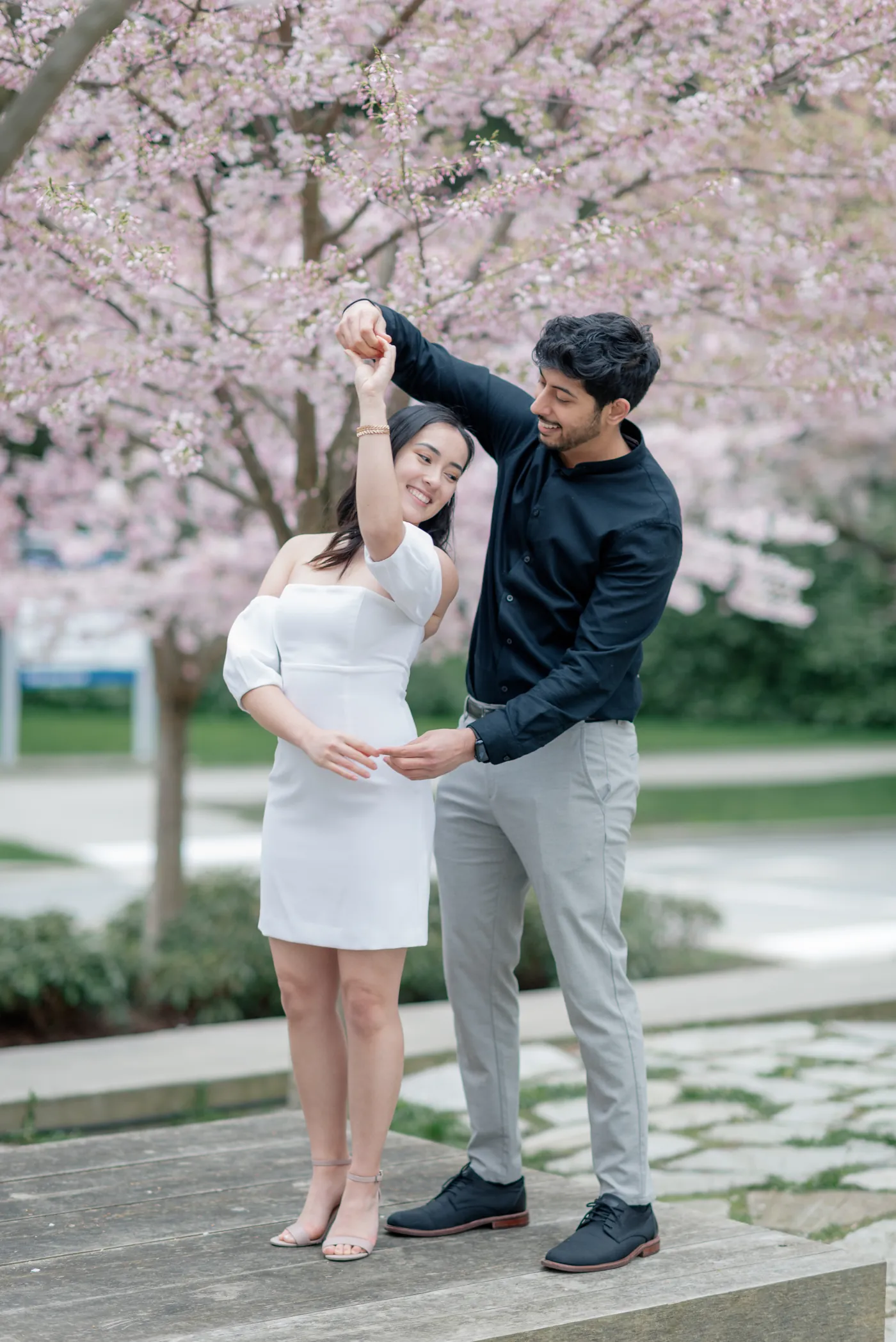 A couple twirls together under a canopy of cherry blossoms. The man, dressed in a black shirt and gray pants, smiles as he lifts his partner’s hand. She wears a white off-shoulder dress and beams with joy.