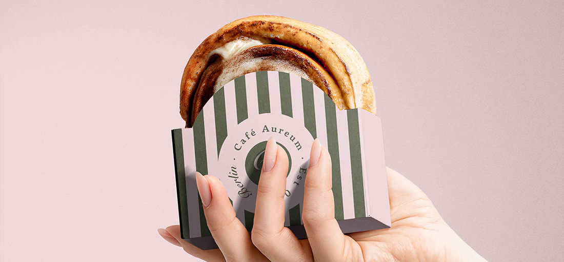 Café Aureum striped sandwich sleeve packaging held in hand, featuring embossed monogram seal — hospitality packaging design by Studio Ruby Signorelli, California.