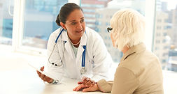 A female doctor in a white coat and stethoscope is sitting at a desk, talking to a senior female patient. The doctor is holding a tablet and showing the patient something on it while also holding her hand in a comforting gesture.