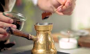 A person wearing a white shirt and black apron is carefully adding a spoonful of coffee grounds to a small copper cezve pot. The cezve pot is sitting on a marble slab and there is a small lit candle beside it.