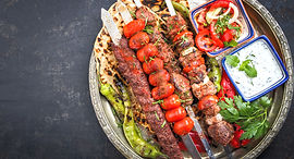 A variety of grilled meat skewers are served on a silver platter with grilled vegetables, fresh flatbread, and two small white bowls holding a yogurt sauce and a tomato relish.
