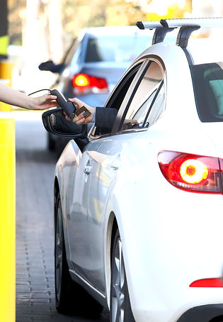A white vehicle is stopped at a toll booth with a window to the attendant's booth on the left. A hand reaches out of the car window to pay the toll.