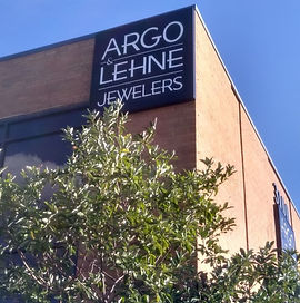 The image is of a brick building with a large sign on it. The sign is black with white lettering and says "Argo & Lehne Jewelers". The building is beside a tree with full green foliage.