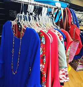Clothes-Mentor-Upper-Arlington-Ohio-Buy-The image displays a clothing rack with various women's clothing items on hangers, including blue tops, floral dresses, and a red blazer, with handbags and a jewelry necklace hanging on the rack as well.