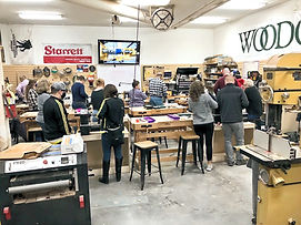 A group of people are working on various woodworking projects in a workshop. Some are using power tools, while others are working on projects at tables.