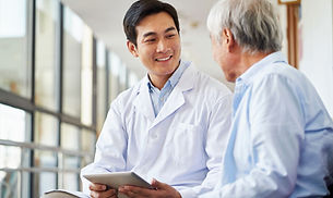 A young Asian male doctor in a white coat sits next to a senior male patient in a blue shirt, holding a tablet and smiling at him.