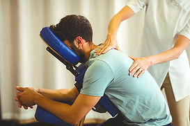 Bearded man in a blue t-shirt sits in a massage chair as a masseuse in white uniform gives him a back massage.