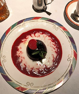 A decadent dessert of a chocolate sphere sits atop a vibrant red raspberry sauce and white cream swirl, garnished with a sprig of mind and a single red  chocolate disc.