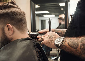 A bearded man sits in a barber chair as a tattooed barber uses an electric razor to trim the back of his hair.